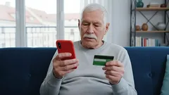 Starší muž platí online mobilem a kreditní kartou, sedí na pohovce doma.