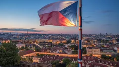 Česká vlajka vlající nad Prahou při západu slunce, panorama města v pozadí.