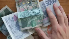 Ruka drží české bankovky s hodnotami 5000 a 2000 korun na dřevěném stole.
